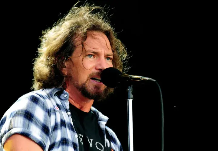 Eddie Vedder, wokalista Pearl Jam, śpiewa do mikrofonu na koncercie. Jego rozwichrzone włosy i charakterystyczna koszula dodają mu rockowego stylu.