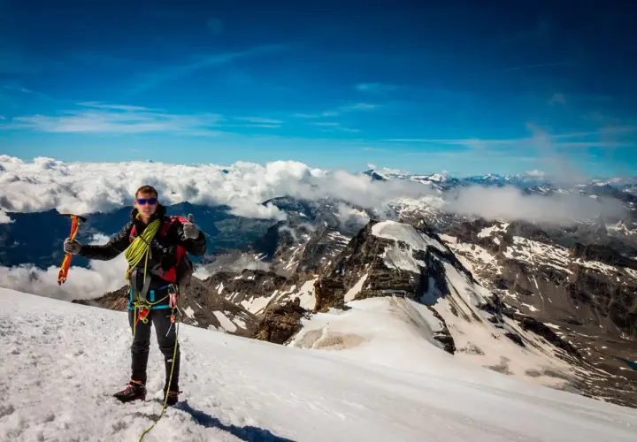 Alpinista en la cima del Gran Paradiso, con piolet y equipo, disfrutando de vistas espectaculares de montañas nevadas y nubes.
