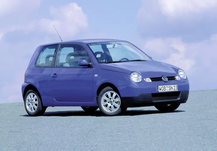 Fioletowy Volkswagen Lupo z tablicą rejestracyjną WOB SN 23 stoi na szarej nawierzchni pod błękitnym niebem z chmurami.