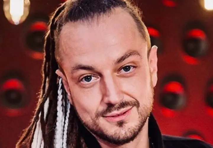 Aleksander Milwiw Baron, w wieku około 40 lat, z dredami i niebieskimi oczami, uśmiecha się na tle czerwonej ściany.