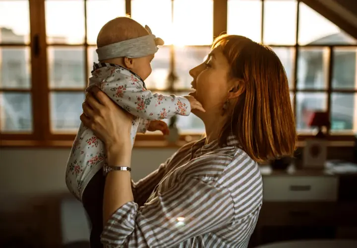 Mama bawi się z dzieckiem, które wykonuje odginanie głowy do tyłu. Ciepłe światło zachodzącego słońca oświetla ich twarze.