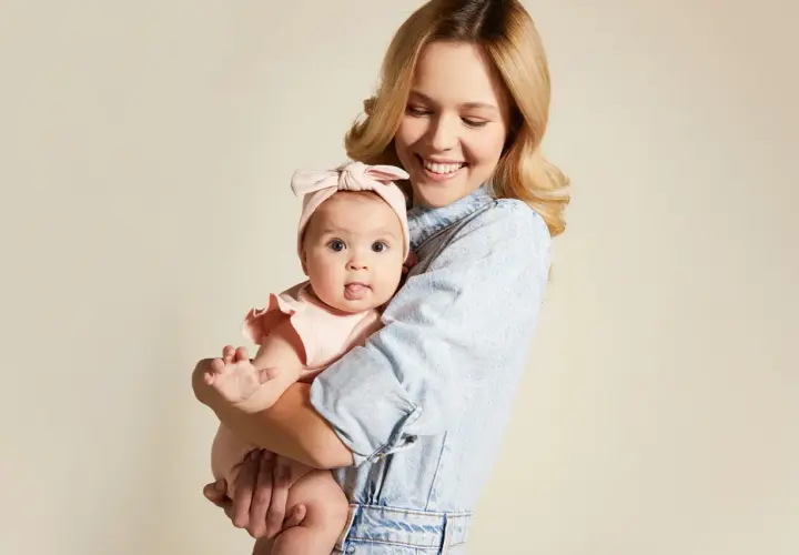 Uśmiechnięta mama tuli niemowlę w różowym body i opasce. Wskazówka, jak ubierać noworodka jesienią, by było mu ciepło i wygodnie.