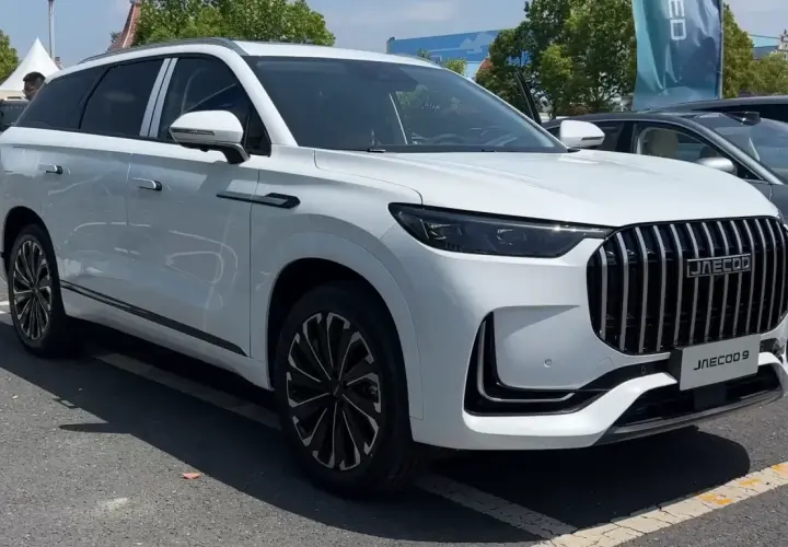 Biały SUV Jaecoo 9 z chromowanym grillem i czarnymi felgami stoi na parkingu.