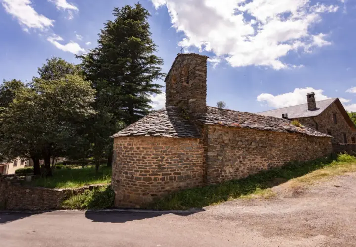 Iglesia románica de piedra con tejado de pizarra en el Ripollès, bajo un cielo azul con nubes algodonosas.