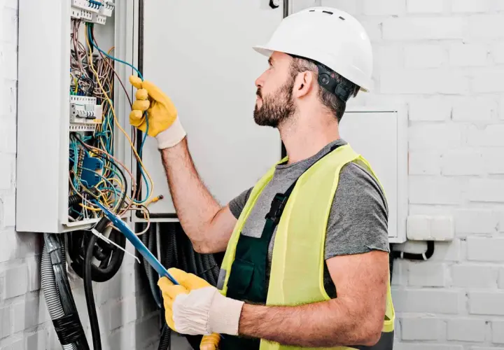 Elektryk w kasku i kamizelce odblaskowej pracuje przy skrzynce z przewodami. Zastanawiasz się, ile kosztuje elektryk?