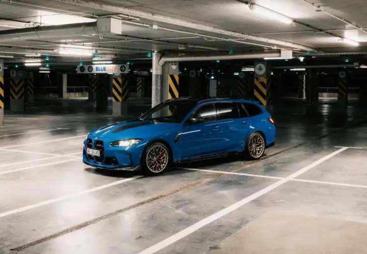 Niebieskie BMW M3 Touring z czarnymi pasami na masce stoi na pustym parkingu podziemnym.