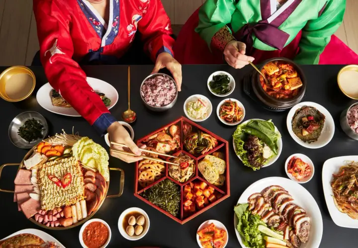 Dwie osoby w tradycyjnych koreańskich strojach jedzą obfity posiłek z wieloma daniami, w tym kimchi.
