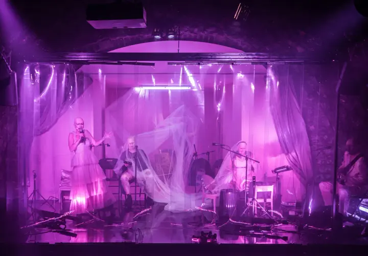 Na scenie teatru ludowego, w fioletowym świetle, artyści prezentują swój repertuar. Wokalistka w zwiewnej sukni i muzycy tworzą magiczną atmosferę.