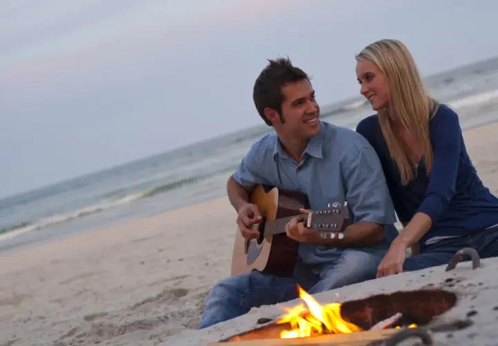 Coppia sulla spiaggia al tramonto, lui suona la chitarra, lei lo guarda sorridendo. Un falò illumina la scena, ma la noia nella coppia è palpabile.