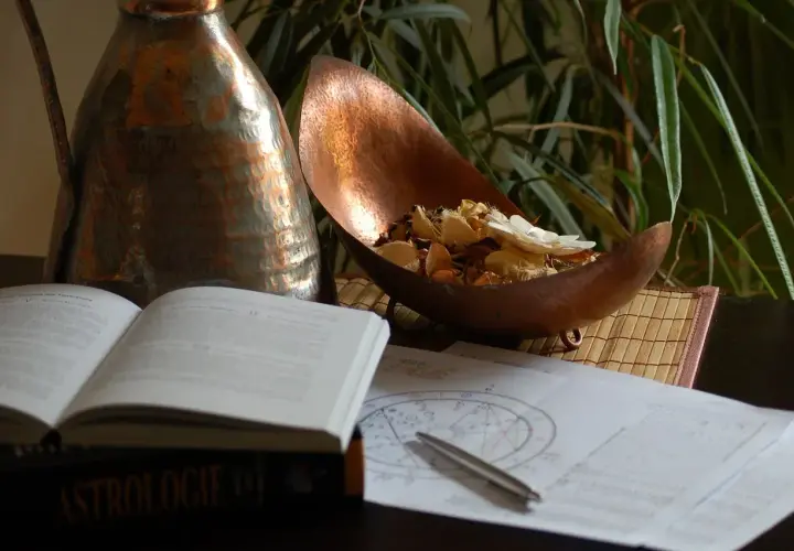 Na stole leży otwarta księga "Astrologie", obok wykresy, długopis i miedziana misa z suszonymi kwiatami. Umiarkowanie tarot uczucia.