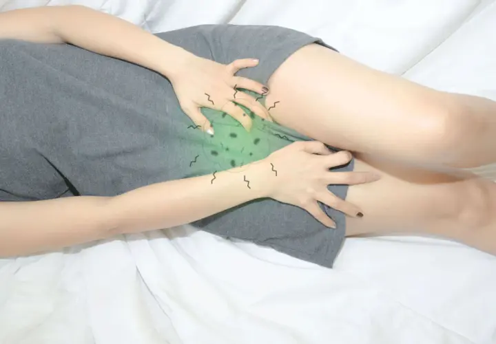 Mujer se rasca la zona íntima, sintiendo molestos picores en la vulva. La imagen muestra su mano sobre la ropa, con un aura verde y garabatos que sugieren irritación.