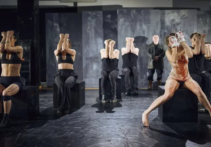 Scena z "Teatru Pieśni Kozła". Aktorzy w mrocznej scenerii, niektórzy z rękami przy twarzy, inni w krwawych strojach.