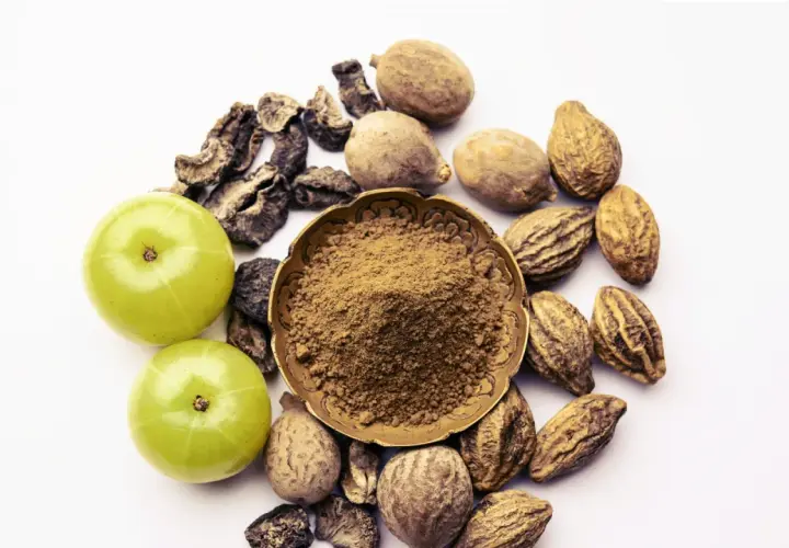 Triphala Pulver in einer Schale, umgeben von Amla-Früchten und getrockneten Haritaki- und Bibhitaki-Samen. Die Triphala Wirkung wird durch diese Zutaten erzielt.