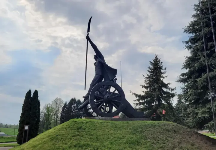 Pomnik Kościuszki z kosą na armacie, symbol zwycięstwa pod Racławicami, na tle pochmurnego nieba i drzew.