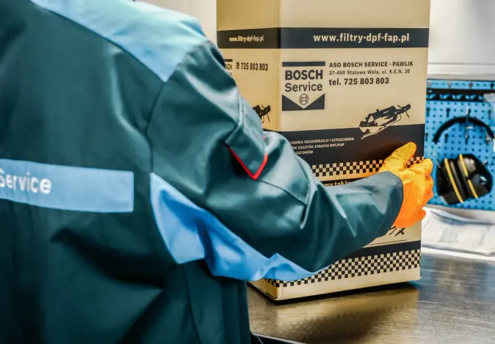 Pracownik w kombinezonie i rękawiczkach trzyma karton z logo Bosch Service.