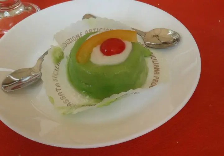 Zielone lody cassate, ozdobione wiśnią i skórką pomarańczy, na białym talerzyku z łyżeczką.