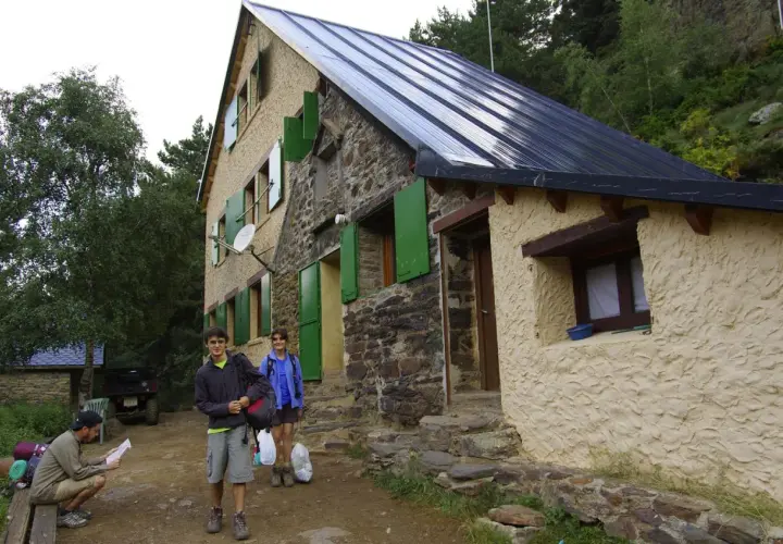 Llegada al refugio de Vallferrera. Dos jóvenes con mochilas y un hombre sentado revisando un mapa esperan en la entrada de piedra y estuco.