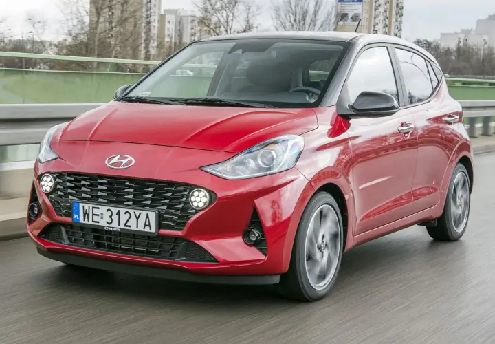 Czerwony Hyundai i10 z czarnym dachem pędzi po drodze. W tle widać budynki i drzewa.