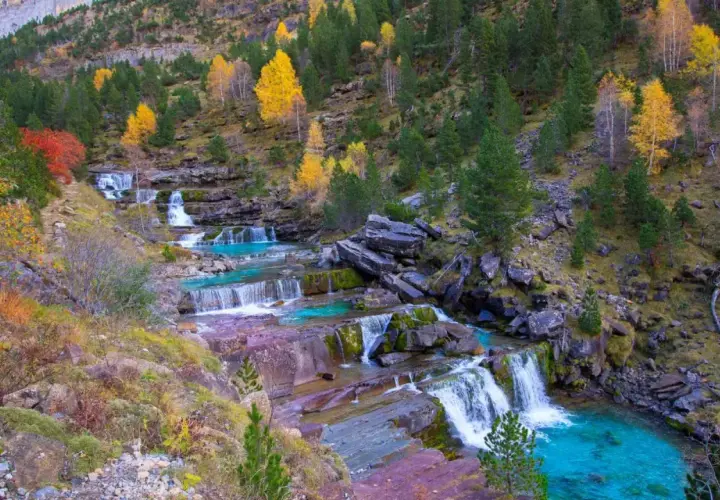 Cascada en las gradas de soaso, con aguas turquesas y rocas rojizas, rodeada de árboles otoñales y pinos verdes.