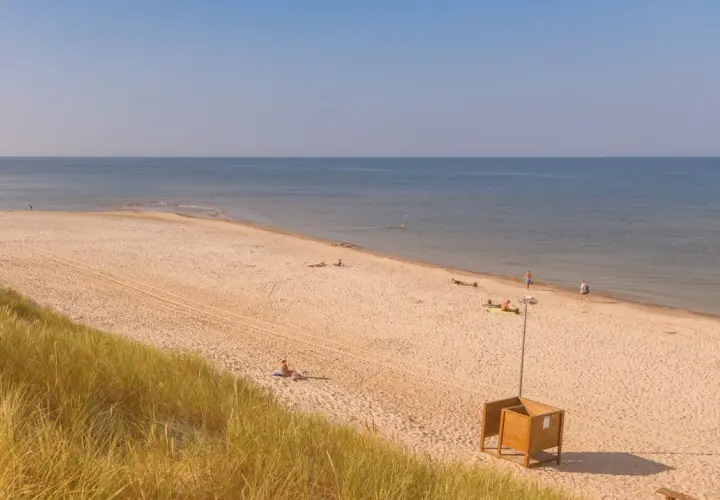 Słoneczna plaża w Kuźnicy nad morzem. Kilka osób odpoczywa na piasku, w oddali widać spokojne morze i błękitne niebo.