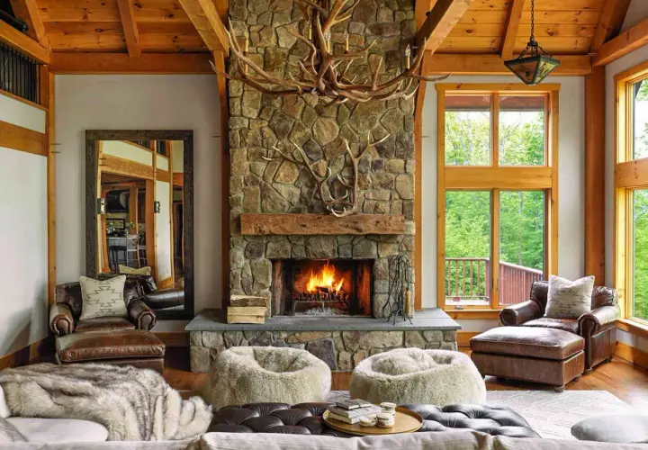 A cozy living room with a stone fireplace, antler chandelier, and leather armchairs embodies traditional rustic interior design.