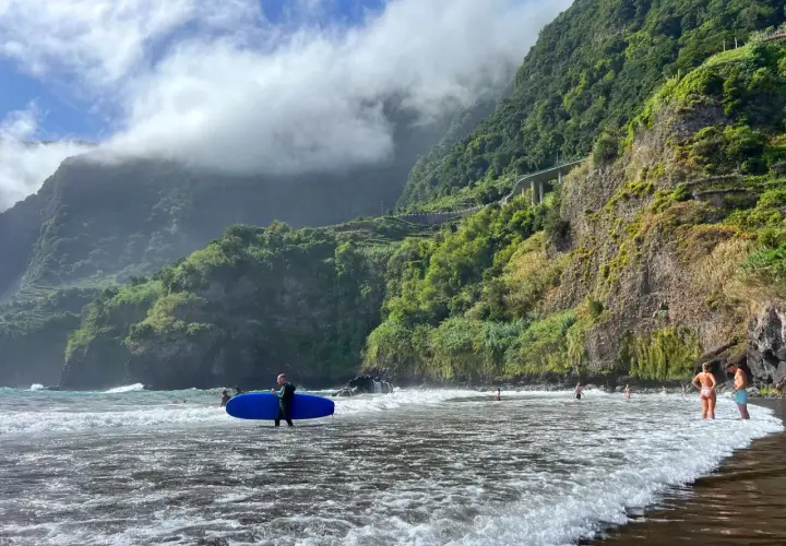 Surfer z niebieską deską na czarnej plaży Madera, w tle zielone klify i chmury.
