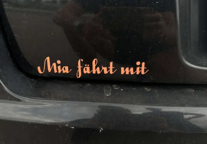 Pomarańczowa naklejka na czarnym lakierze samochodu z napisem "Mia fährt mit". Chcesz wiedzieć, jak usunąć naklejki z samochodu?