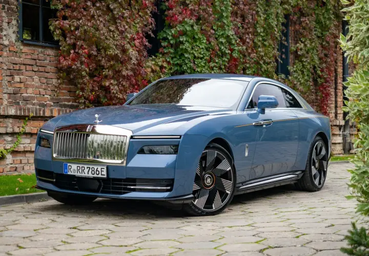 Niebieski Rolls-Royce Spectre, luksusowy samochód elektryczny, zaparkowany przed ceglanym budynkiem porośniętym jesiennymi liśćmi.