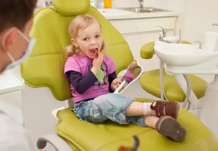 Mała dziewczynka z lusterkiem sprawdza swoje zdrowe zęby mleczne podczas wizyty u dentysty.