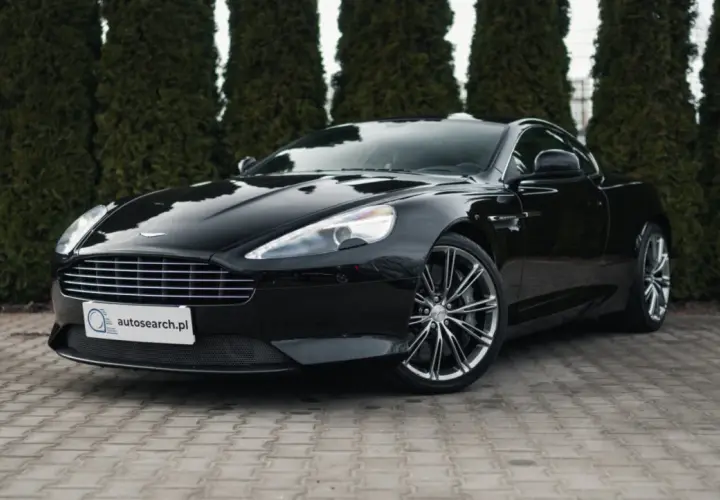 Czarny Aston Martin DB9 lśni na tle zielonych żywotników. Elegancki, sportowy samochód z charakterystycznym grillem i felgami.