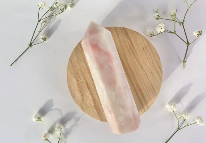 Ein polierter rosa Opal-Kristall liegt auf einem Holzsockel, umgeben von zarten weißen Blüten. Die rosa Opal Wirkung wird oft mit Liebe und Heilung assoziiert.