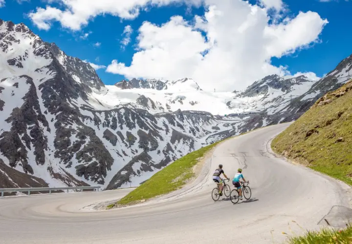Two cyclists enjoy an epic adventure on a winding road, part of amazing austria bike tours, with snow-capped mountains in the background.
