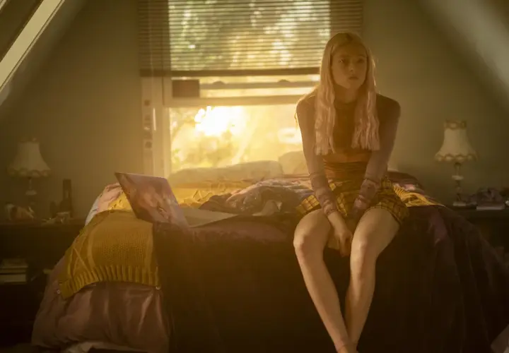 Una ragazza bionda con un vestito a quadri siede sul letto, illuminata dalla luce del tramonto. Una scena che fa pensare a una recensione di Euphoria.