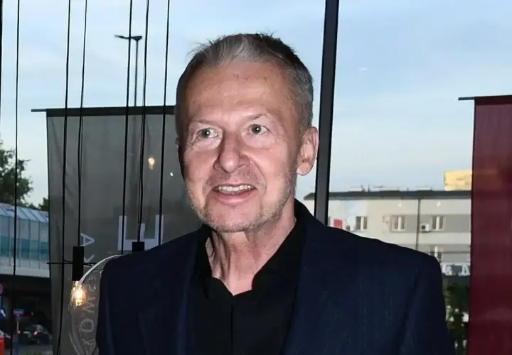 Bogusław Linda w granatowej marynarce i czarnej koszuli, uśmiecha się.