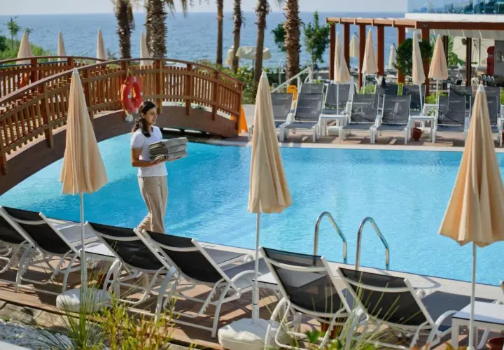 Pracownica hotelu niesie ręczniki przy basenie w Avsallar. W tle widać morze i palmy.