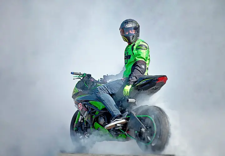 Motociclista con casco y traje verde realiza un caballito, generando humo. La moto, con detalles verdes, parece lista para una demostración de homologación e24.