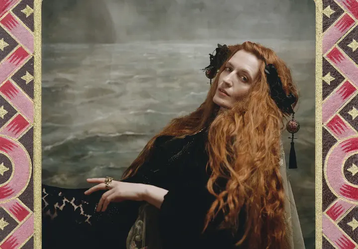 Florence Welch, jak królowa, z rudymi włosami i w czarnej sukni, na tle morskiego krajobrazu.