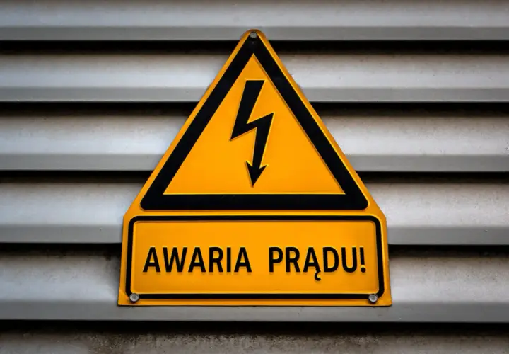 Żółty trójkąt z symbolem pioruna i tabliczka "AWARIA PRĄDU!" na szarej, metalowej powierzchni.