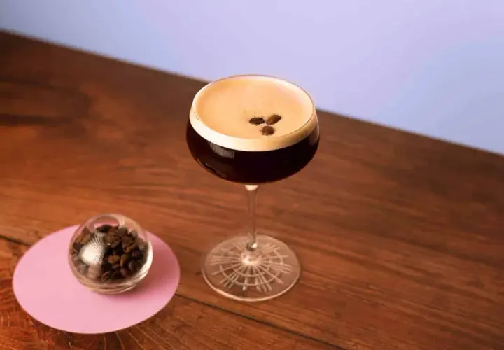 Espresso Martini with coffee beans on top, next to a small glass of coffee beans on a pink coaster.