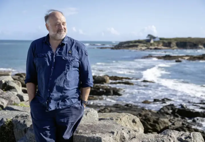 Ein Mann in blauer Kleidung steht am Meer. Die bannalec reihenfolge der Wellen spiegelt sich in seiner entspannten Haltung wider.