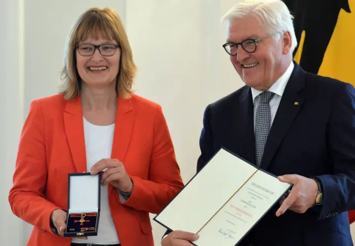 Frau mit rotem Blazer erhält das Bundesverdienstkreuz. Präsident Steinmeier überreicht die Auszeichnung und Urkunde.