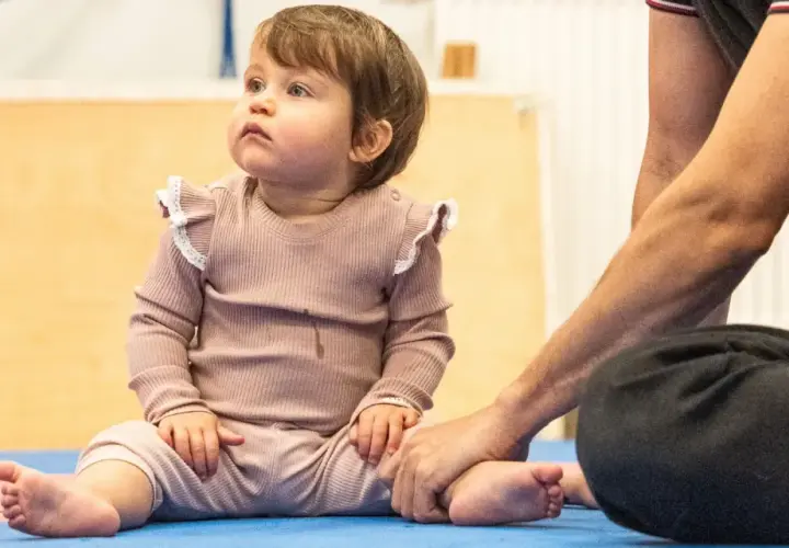 Małe dziecko siedzi na macie, a dorosły trzyma je za stopę. Czy 5 miesięczne dziecko można sadzać na kolanach?