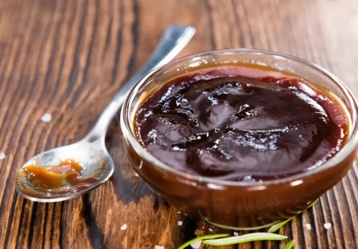 Un cuenco de cristal lleno de deliciosa salsa barbacoa casera, con una cuchara al lado y romero fresco.