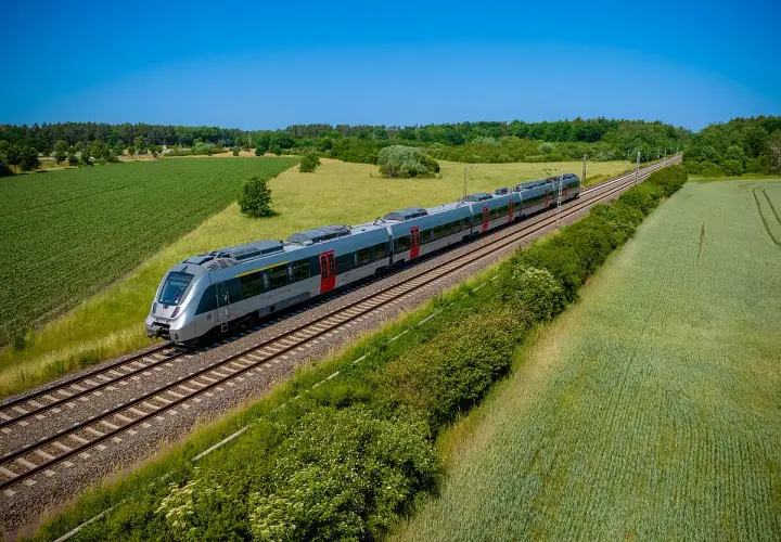 Moderner Triebzug fährt auf einer **railway deutsch** durch grüne Felder und Wälder unter blauem Himmel.