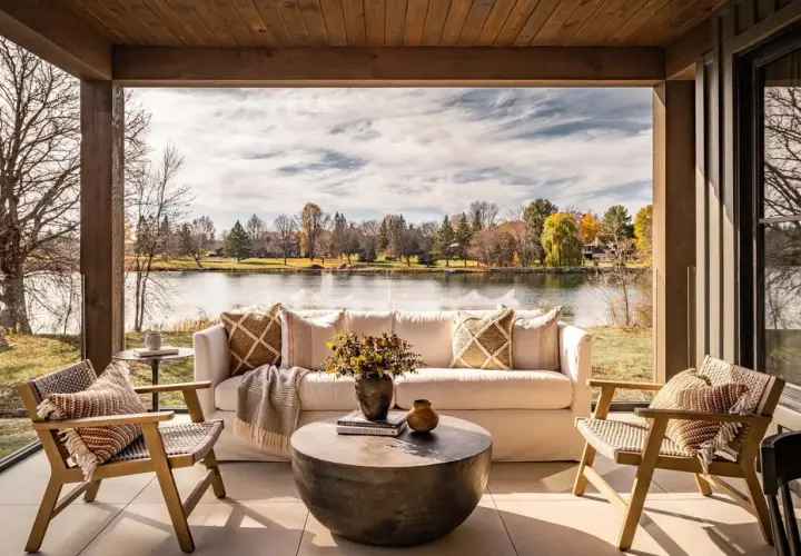 Cozy lakehouse interior design with a white sofa, chairs, and coffee table on a covered patio overlooking a serene lake and autumn foliage.