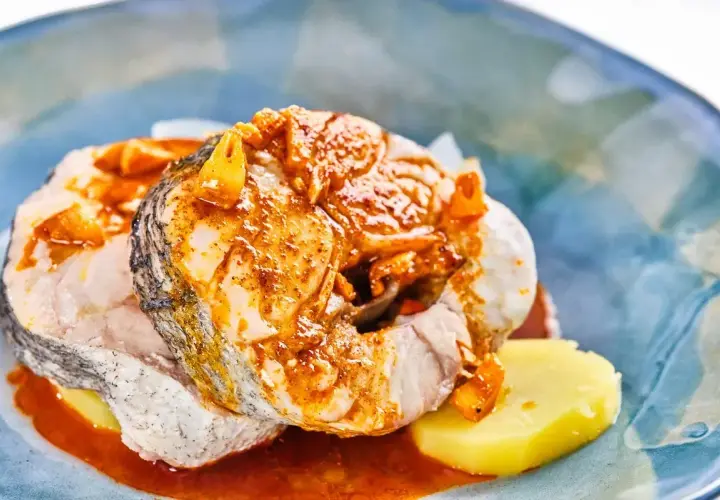 Rodajas de pescado blanco cubiertas con una sabrosa salsa gallega, servidas sobre patatas cocidas en un plato azul.