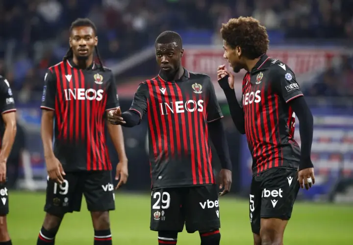Piłkarze OGC Nice w czarno-czerwonych strojach dyskutują na boisku. Jeden z nich, z numerem 29, wskazuje palcem.