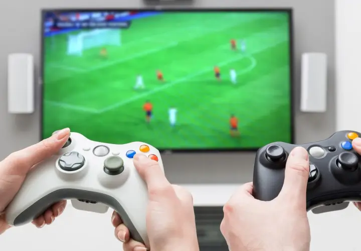 Dwie osoby grają w gry sportowe na konsoli, trzymając kontrolery przed ekranem telewizora z boiskiem piłkarskim.