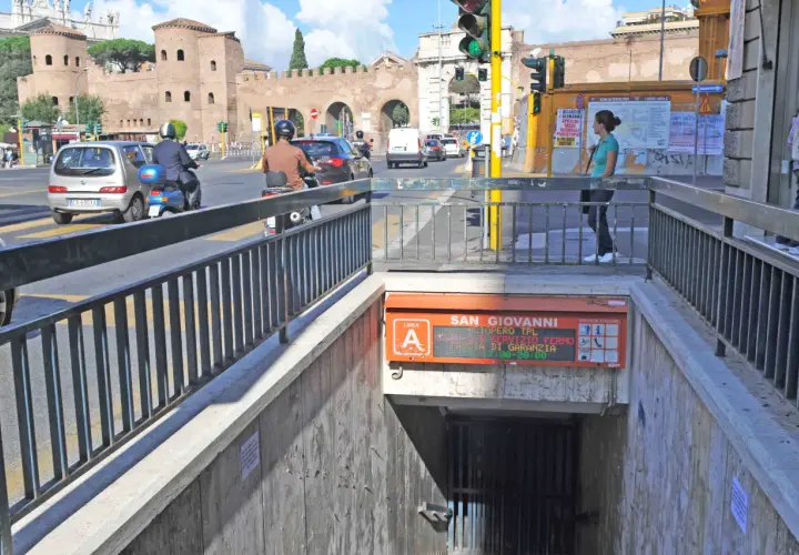 Ingresso metro San Giovanni, collegamento metro A. Traffico intenso con auto e scooter, sullo sfondo le antiche mura romane.