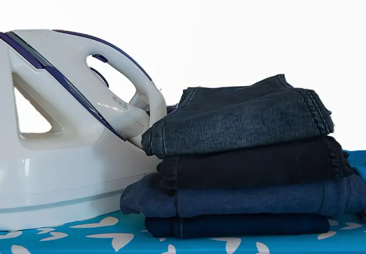 Fer à repasser et pile de vêtements sombres sur une planche à repasser. Apprenez comment repasser du polyester pour un résultat impeccable.